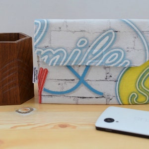 K&ouml;nnte beinhalten: Eine wei&szlig;e Handyh&uuml;lle aus Stoff mit einem Ziegelmauermuster und dem Wort "smile" in blauen Neonbuchstaben. Die H&uuml;lle hat au&szlig;erdem ein gelbes und gr&uuml;nes Neon-Design.