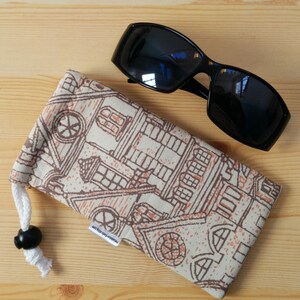 May include: A black sunglasses case with a white background and a brown and orange pattern of houses. The case has a drawstring closure and a black toggle.
