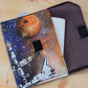 May include: A brown fabric case with a zipper closure and a space-themed print featuring a planet, a spaceship, and a space station. The case is open to reveal a white tablet inside.