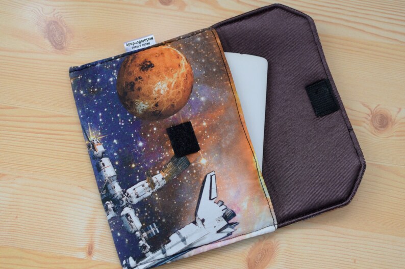 May include: A brown fabric case with a zipper closure and a space-themed print featuring a planet, a spaceship, and a space station. The case is open to reveal a white tablet inside.