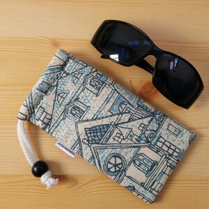 May include: A white and blue patterned fabric sunglasses case with a white drawstring and a black bead. The case is lying on a wooden surface next to a pair of black sunglasses.