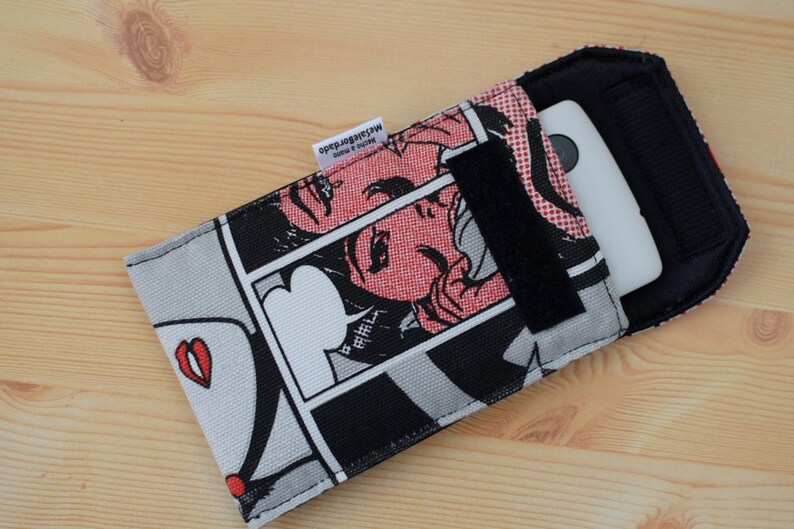Pu&ograve; includere: Una custodia per telefono in bianco e nero con un design in stile fumetto che presenta una donna con labbra rosse e un fumetto che dice "B-R-I-L-L". La custodia ha una patta nera con chiusura a velcro.