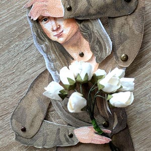 May include: A paper cutout of a woman in a brown dress, holding a bouquet of white flowers. The woman's arm is raised to her forehead. The cutout is attached with small brass rivets. The background is a light brown wood grain.