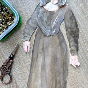 May include: Watercolor paper doll of the Mona Lisa in a brown dress. The paper doll is on a wooden surface with a green container of gold tacks, a watercolor palette, and decorative scissors.