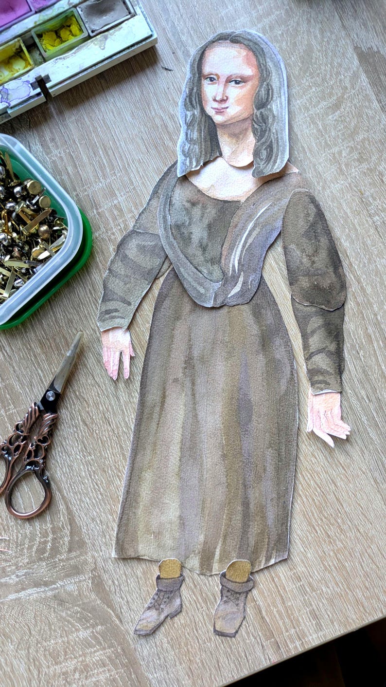 May include: Watercolor paper doll of the Mona Lisa in a brown dress. The paper doll is on a wooden surface with a green container of gold tacks, a watercolor palette, and decorative scissors.