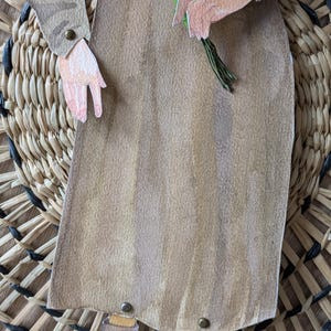 May include: A paper cutout of a person in a long brown dress holding a bouquet of white flowers. The figure is set against a woven background. The person's arms and legs are visible, with details like buttons and socks.