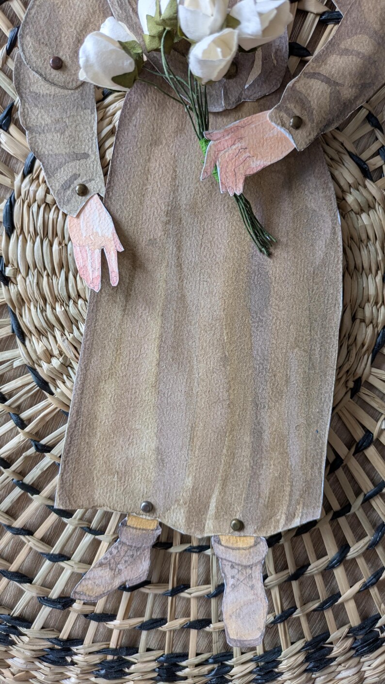 May include: A paper cutout of a person in a long brown dress holding a bouquet of white flowers. The figure is set against a woven background. The person's arms and legs are visible, with details like buttons and socks.