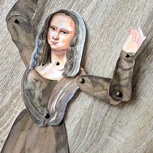 May include: A watercolor paper doll of the Mona Lisa with articulated arms. The doll is dressed in a brown gown and has long gray hair. The arms are jointed with small brass rivets, allowing for movement.