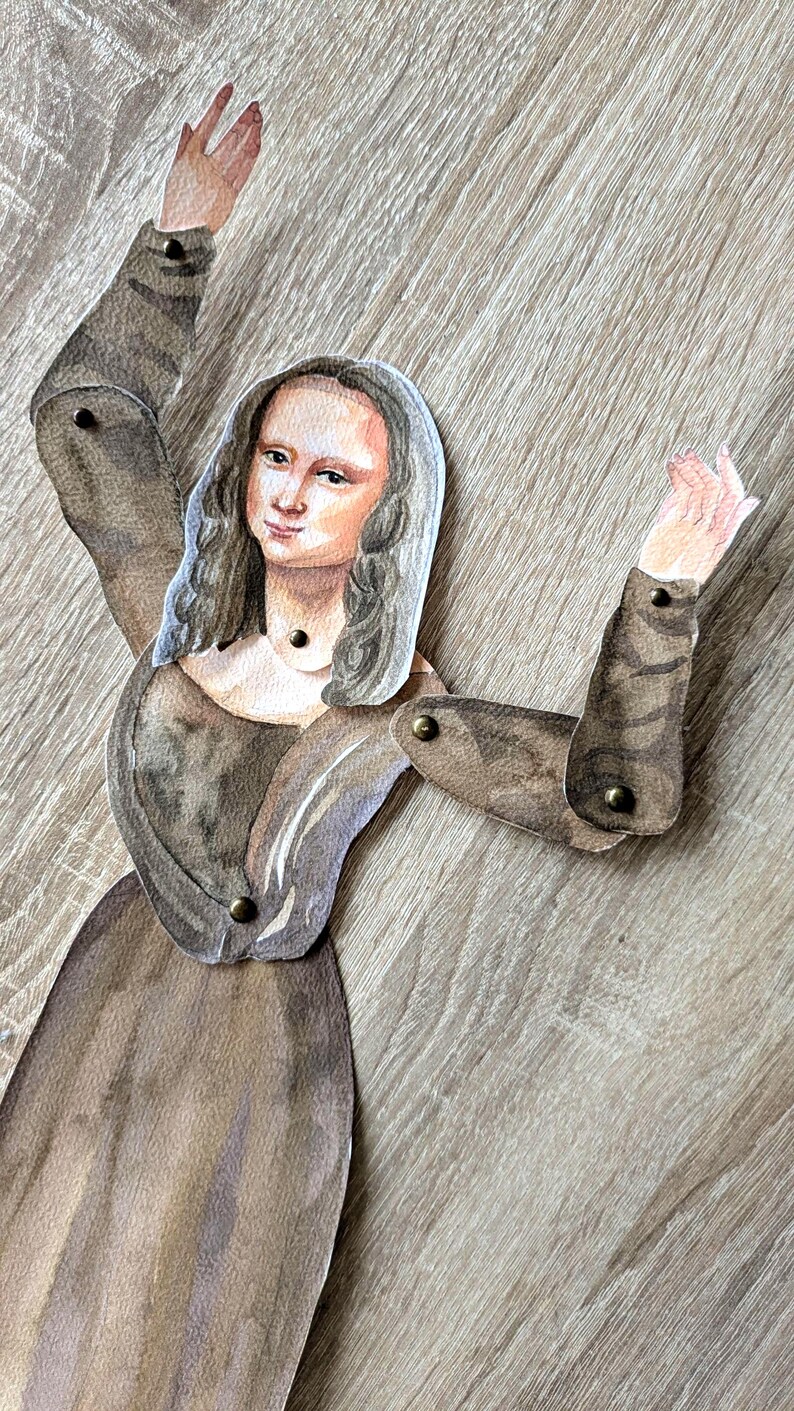 May include: A watercolor paper doll of the Mona Lisa with articulated arms. The doll is dressed in a brown gown and has long gray hair. The arms are jointed with small brass rivets, allowing for movement.