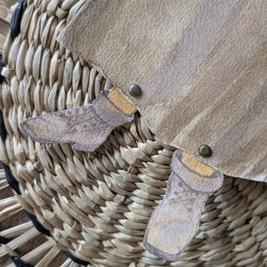 May include: Close-up of a woven basket with a paper cutout design. The paper is tan with brown accents and two small paper cutouts of boots. The basket is made of natural fibers.