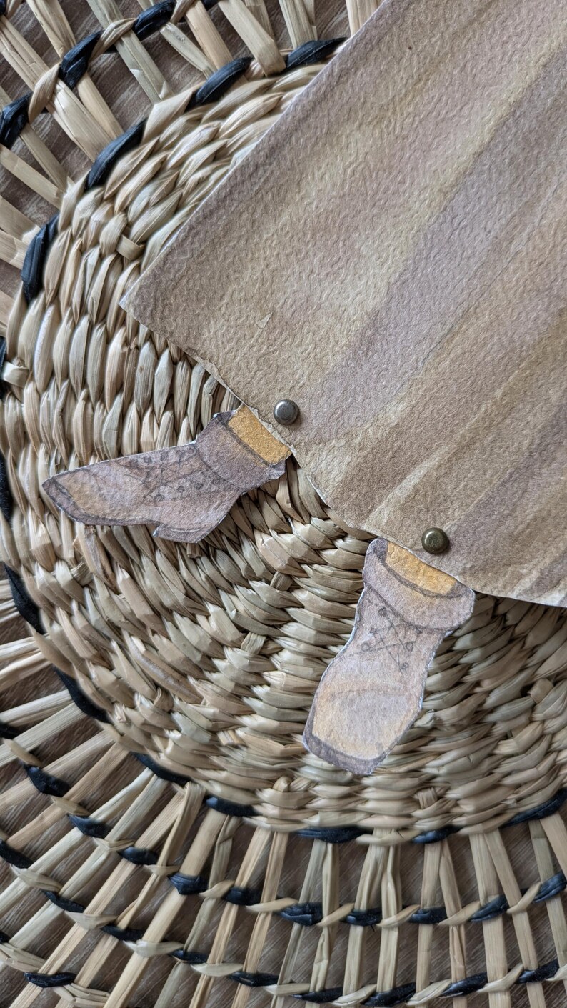 May include: Close-up of a woven basket with a paper cutout design. The paper is tan with brown accents and two small paper cutouts of boots. The basket is made of natural fibers.