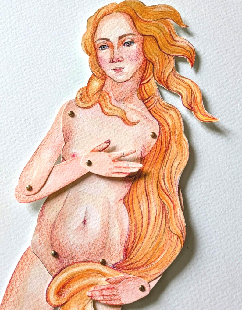 A paper doll of a woman with long flowing red hair. The doll is made of paper and has been painted with watercolor and colored pencils. The doll has a light pink skin tone and is wearing a light pink dress. The doll's hair is long and flowing and is painted in shades of red, orange, and yellow. The doll's eyes are blue and her lips are red. The doll is standing on a white background.