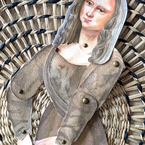 May include: A paper cutout of the Mona Lisa, with articulated arms, is positioned over a woven basket. The artwork features a brown dress and long, wavy hair. The basket has a circular design with natural and black woven elements.