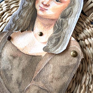 May include: A paper cutout of the Mona Lisa, featuring a watercolor painting style. The figure is dressed in a brown gown and has brass fasteners at the joints. The background is a woven, natural-toned material.