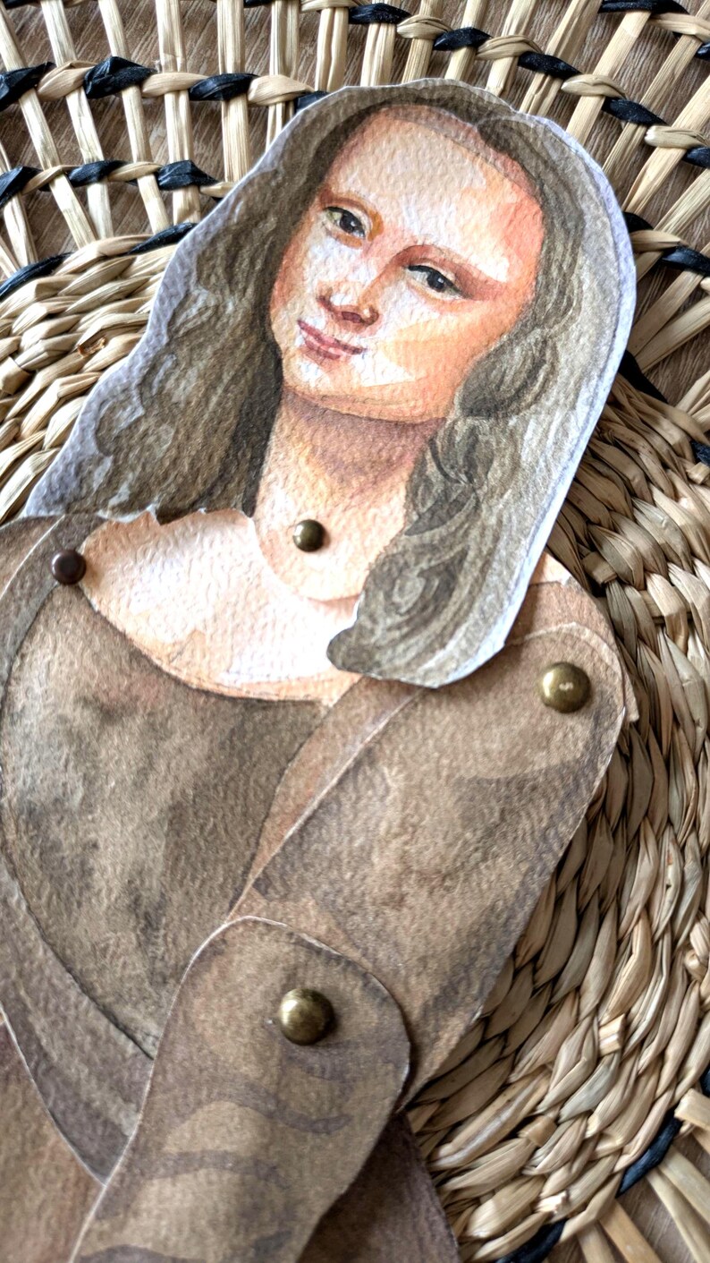 May include: A paper cutout of the Mona Lisa, featuring a watercolor painting style. The figure is dressed in a brown gown and has brass fasteners at the joints. The background is a woven, natural-toned material.