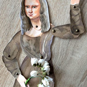 May include: A paper cutout of the Mona Lisa in a brown dress, holding white flowers. The figure has articulated arms and is attached with small metal rivets. The background is a light brown wood grain.