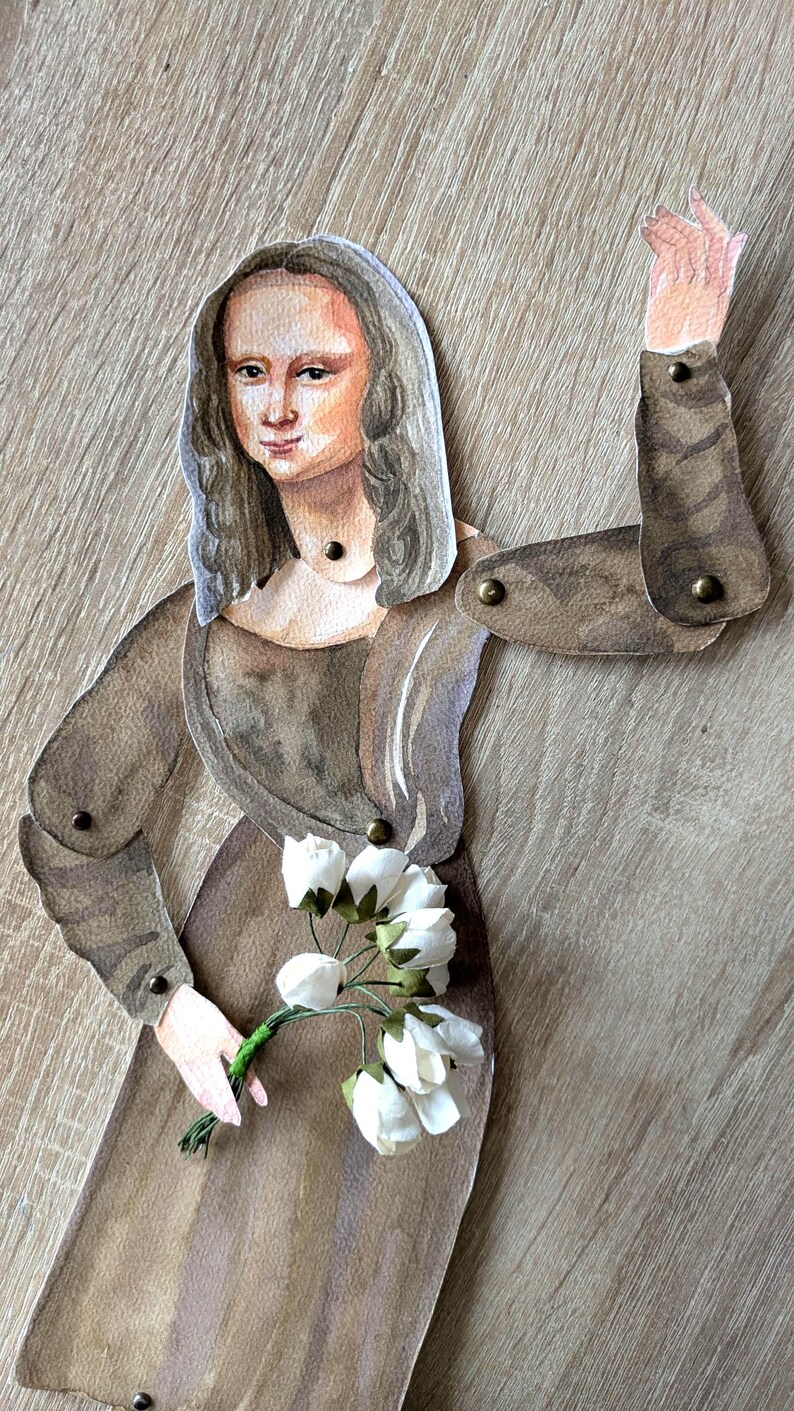 May include: A paper cutout of the Mona Lisa in a brown dress, holding white flowers. The figure has articulated arms and is attached with small metal rivets. The background is a light brown wood grain.