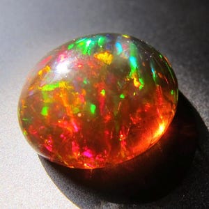 Op de afbeelding: Een gepolijste, ovaalvormige zwarte opaal edelsteen. De opaal vertoont een levendig kleurenspel, met flitsen van rood, groen, geel en blauw. De edelsteen is geplaatst tegen een donkere achtergrond, wat de iriserende kwaliteiten benadrukt.