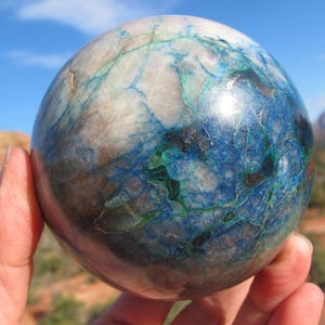May include: A polished sphere with a marbled appearance, featuring a blend of blue, green, and white hues. The stone ball is held in a hand, with a backdrop of a blue sky and distant mountains.