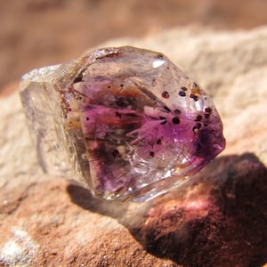 May include: A clear, faceted gemstone with a vibrant purple interior and dark inclusions. The crystal is resting on a rough, reddish-brown stone surface, with sunlight casting a shadow. The gemstone's unique color and clarity are highlighted.