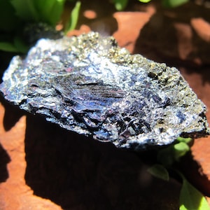 Pode incluir: Uma aproximação de um espécime mineral com brilho metálico. A rocha tem uma aparência escura e em camadas com tons iridescentes de azul, roxo e dourado. O espécime é colocado contra um fundo natural de folhas verdes e uma superfície marrom.