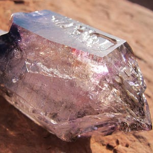 Può includere: Un grande cristallo di ametista sfaccettato con una parte superiore rettangolare lucida e un corpo ruvido e strutturato. Il cristallo mostra sfumature di viola, lavanda e trasparente, con la luce che si riflette sulle sue superfici. È una gemma naturale.