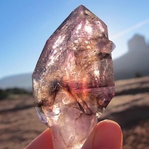 Può includere: Un cristallo di ametista traslucido con una punta appuntita, che mostra sfumature di viola, marrone e trasparente. Il cristallo è tenuto contro uno sfondo di cielo soleggiato e un paesaggio sfocato.