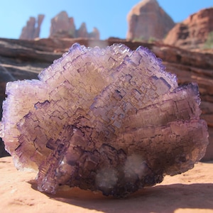 May include: A large, purple and clear fluorite crystal with a layered, geometric structure. The crystal is sitting on a red rock surface.