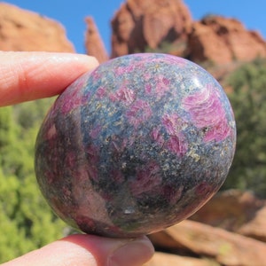 Puede incluir: Una esfera de piedra preciosa lisa y pulida, de color gris oscuro y rosa, con un patrón de remolino. La esfera está siendo sostenida en una mano.