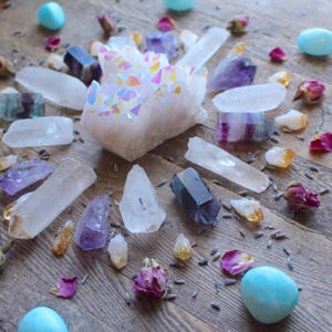May include: A crystal grid with a large iridescent quartz cluster in the center, surrounded by smaller clear quartz crystals, amethyst, fluorite, and turquoise stones. The grid is laid out on a wooden surface with dried flowers and petals scattered around.