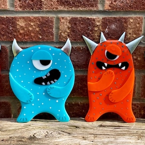 Könnte beinhalten: Vier farbenfrohe Monster-Dekorationen. Die Monster sind rot, blau, orange und weiß, jedes mit einzigartigen Merkmalen wie Hörnern, Augen und Gesichtsausdrücken. Sie sind in einer Reihe vor einer Ziegelmauer angeordnet.