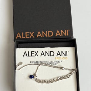 May include: A silver-toned bracelet with a chain link design and a small blue eye charm, presented in a black box. The box lid displays the brand name "ALEX AND ANI" in gold lettering. The bracelet is part of the "PROVIDENCE COLLECTION".