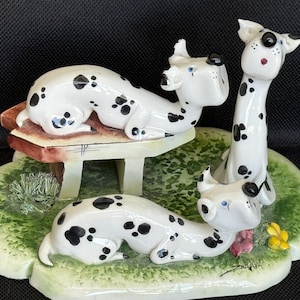 May include: A ceramic figurine featuring three dalmatian dogs. Two dogs are lying down, one on a small bench. The dogs are white with black spots, and the base is green with painted grass and flowers.