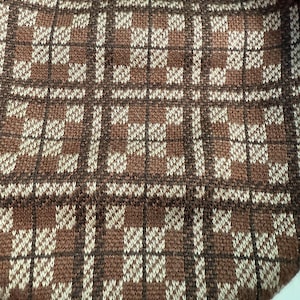 May include: Close-up of a brown and cream-colored knitted fabric with a plaid pattern. The design features a grid of dark brown lines with cream-colored and brown squares. The fabric has a textured appearance.