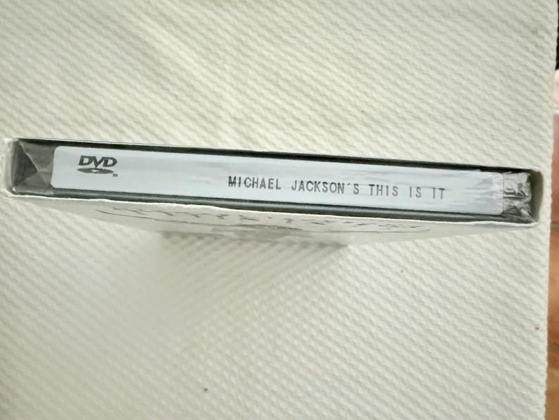 May include: A sealed DVD case for Michael Jackson's This Is It. The spine is white with black text reading DVD and Michael Jackson's This Is It. The case is wrapped in clear plastic film, showing the side profile of the rectangular media product.