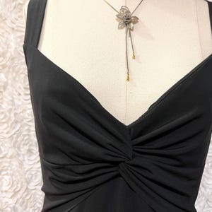 May include: A black dress with a V-neckline and a gathered knot detail at the waist. The dress has wide straps and is displayed on a mannequin. A silver-toned necklace with a flower pendant and gold accents hangs on the mannequin.