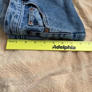 May include: Folded blue denim jeans with copper rivets and a yellow ruler with the word "Adelphia" printed on it. The jeans are folded over, showing the front pockets and belt loops. The fabric is a medium wash denim.