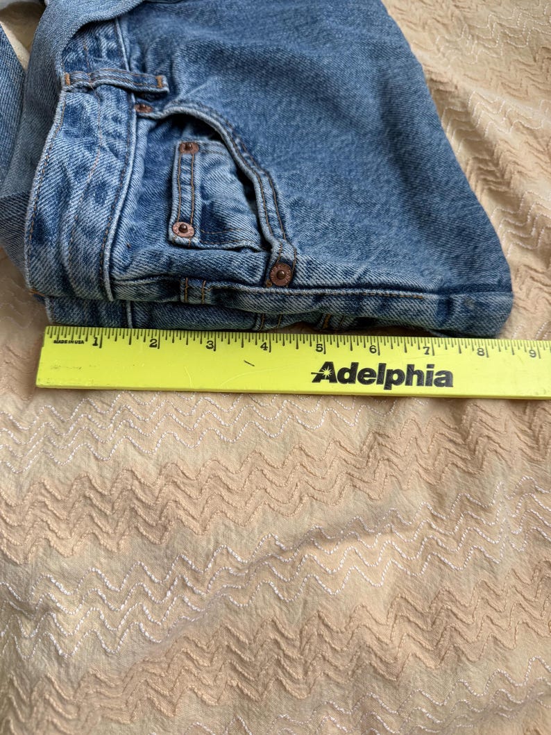 May include: Folded blue denim jeans with copper rivets and a yellow ruler with the word "Adelphia" printed on it. The jeans are folded over, showing the front pockets and belt loops. The fabric is a medium wash denim.
