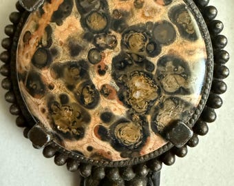 May include: A round bolo tie pendant with a speckled, patterned stone in shades of brown, tan, and black. The stone is set in a dark metal frame with a beaded edge and square corner accents. The braided leather cord is black.
