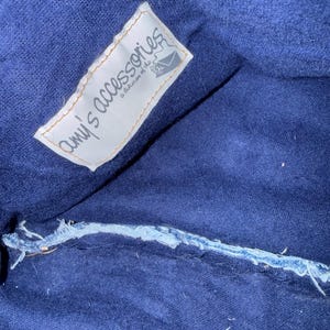May include: Close-up of a dark blue fabric with a white label that reads "amy's accessories a division of the..." The label has a torn edge and a small illustration of an envelope. The fabric has a visible seam.