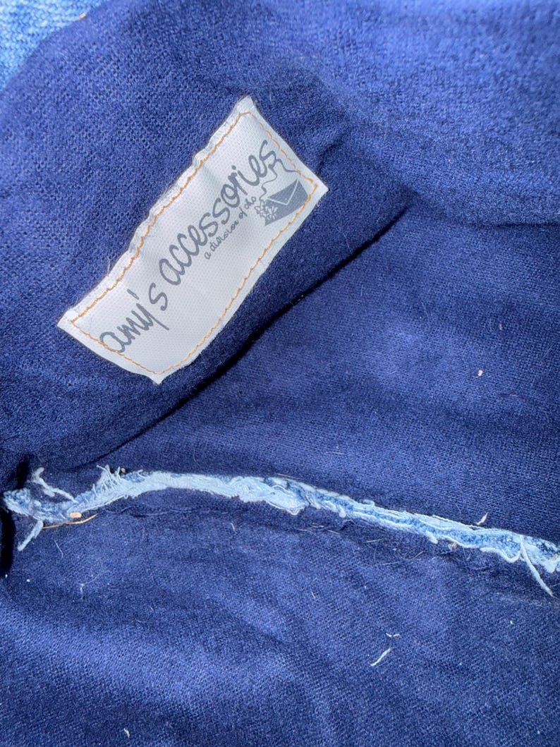 May include: Close-up of a dark blue fabric with a white label that reads "amy's accessories a division of the..." The label has a torn edge and a small illustration of an envelope. The fabric has a visible seam.
