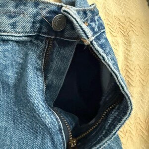 May include: Close-up of light blue denim jeans with a partially unzipped brass zipper. The jeans have a button closure at the waistband and a visible pocket lining. The fabric shows a classic denim texture.