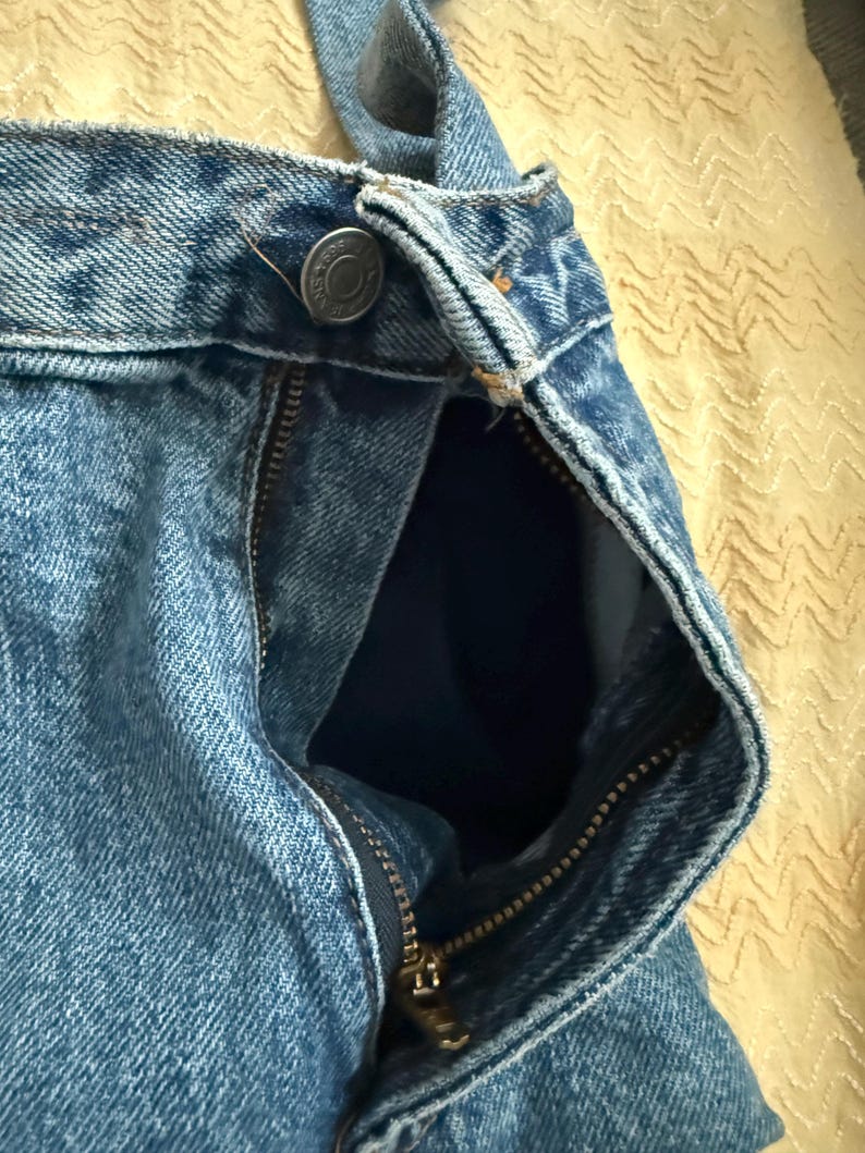 May include: Close-up of light blue denim jeans with a partially unzipped brass zipper. The jeans have a button closure at the waistband and a visible pocket lining. The fabric shows a classic denim texture.