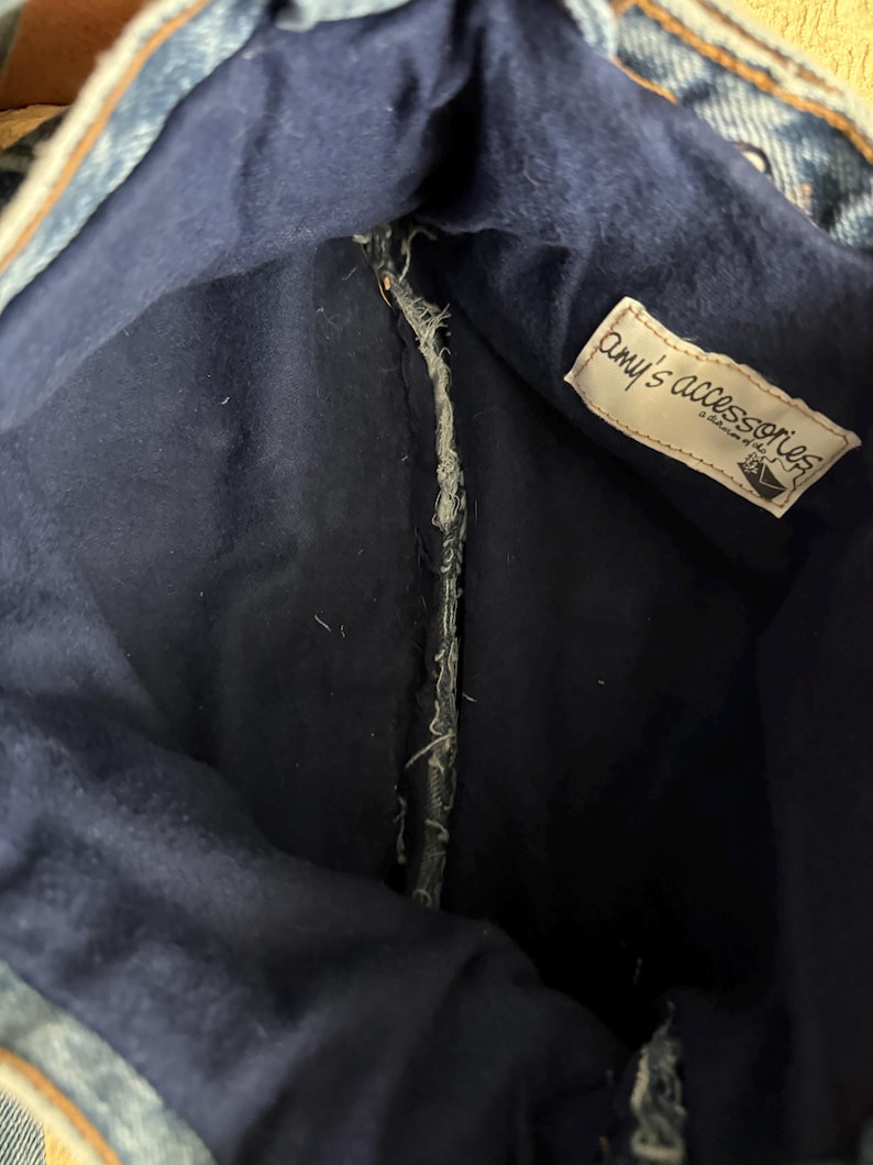 May include: Inside view of a denim bag with a dark blue interior. The bag's interior features a tag that reads "amy's accessories". The bag is made from recycled denim material.