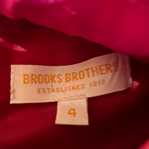 Puede incluir: Primer plano de una etiqueta de Brooks Brothers, establecida en 1818. La etiqueta es rectangular con esquinas redondeadas y presenta el nombre de la marca en una fuente serif. Debajo del nombre de la marca est&aacute; el texto "ESTABLISHED 1818". Una etiqueta de talla 4 est&aacute; debajo. El fondo es una tela rosa brillante.