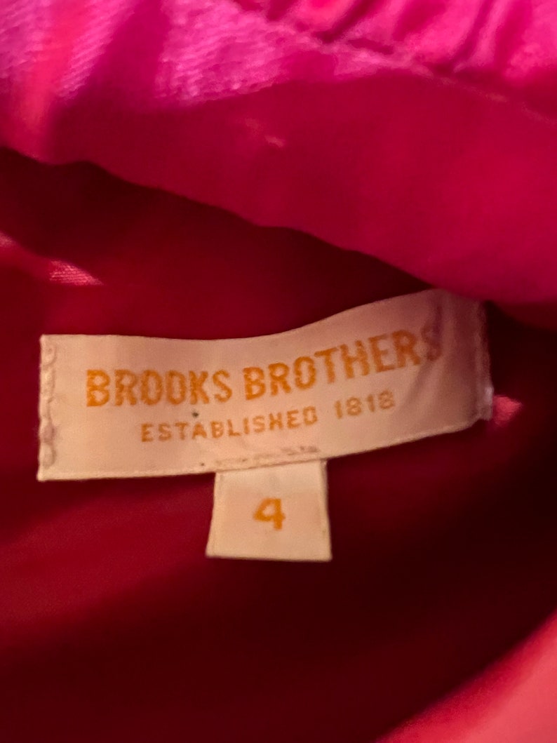 Puede incluir: Primer plano de una etiqueta de Brooks Brothers, establecida en 1818. La etiqueta es rectangular con esquinas redondeadas y presenta el nombre de la marca en una fuente serif. Debajo del nombre de la marca est&aacute; el texto "ESTABLISHED 1818". Una etiqueta de talla 4 est&aacute; debajo. El fondo es una tela rosa brillante.