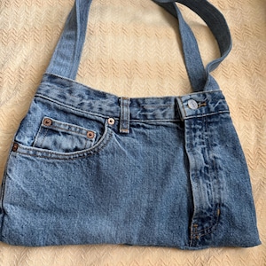 May include: A blue denim handbag with a shoulder strap. The bag is constructed from repurposed jeans, featuring a front pocket, belt loops, and a button closure. The denim has a faded, worn appearance.