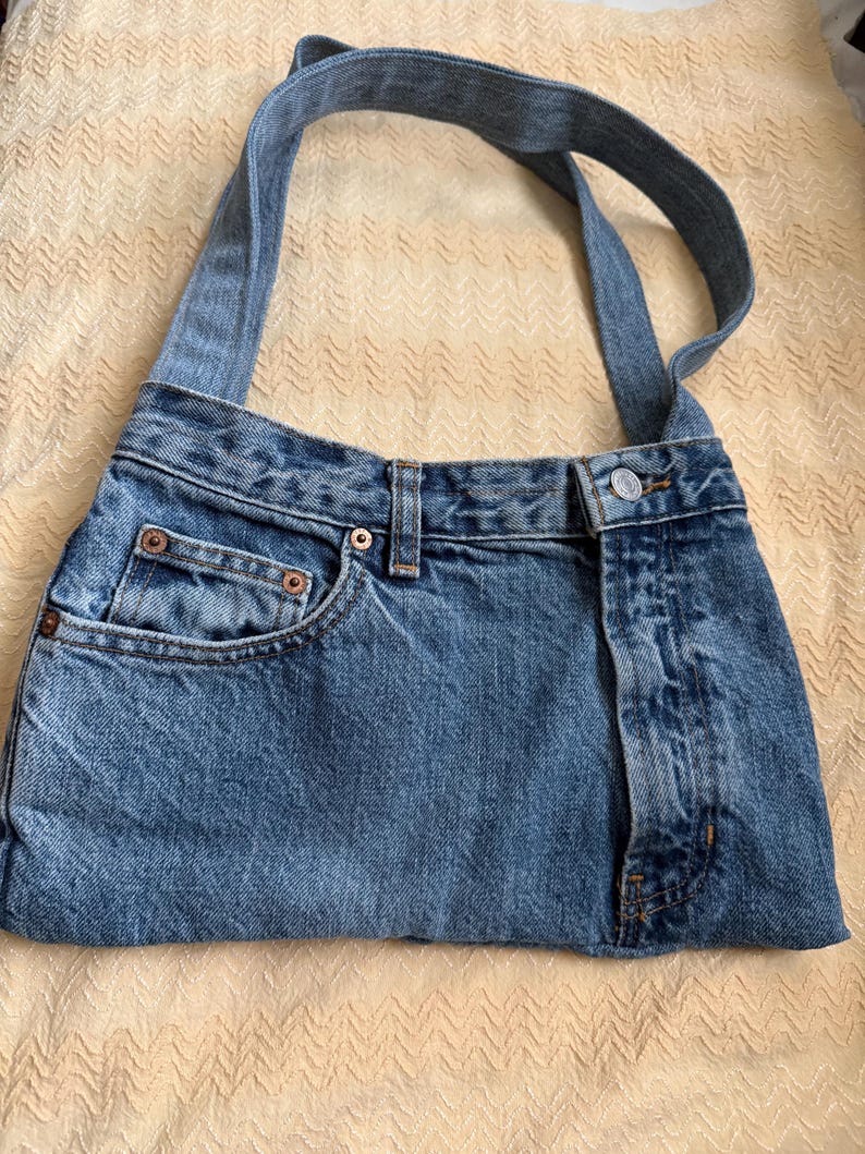 May include: A blue denim handbag with a shoulder strap. The bag is constructed from repurposed jeans, featuring a front pocket, belt loops, and a button closure. The denim has a faded, worn appearance.