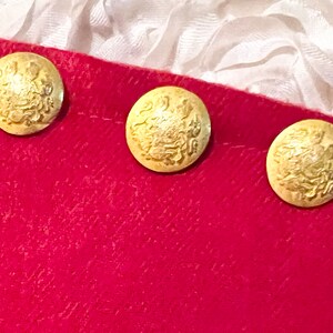 Puede incluir: Tres botones dorados con un dise&ntilde;o en relieve est&aacute;n dispuestos en fila sobre una tela roja. Los botones tienen un patr&oacute;n detallado y ornamentado. El fondo incluye tela blanca.