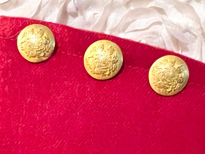 Puede incluir: Tres botones dorados con un dise&ntilde;o en relieve est&aacute;n dispuestos en fila sobre una tela roja. Los botones tienen un patr&oacute;n detallado y ornamentado. El fondo incluye tela blanca.